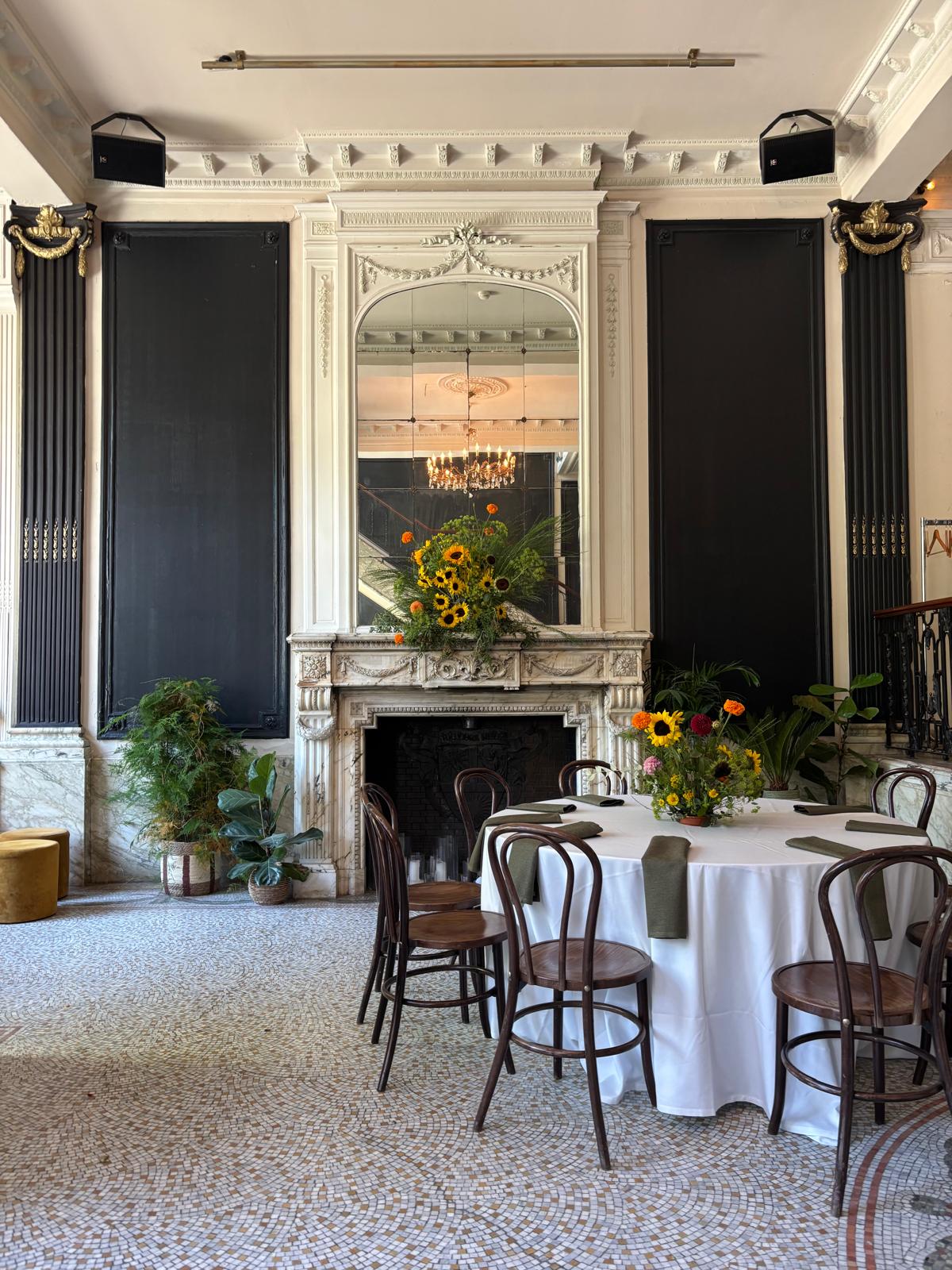 Feestzaal met een gedekte ronde tafel en een groot bloemstuk met zonnebloemen op de marmeren schouw